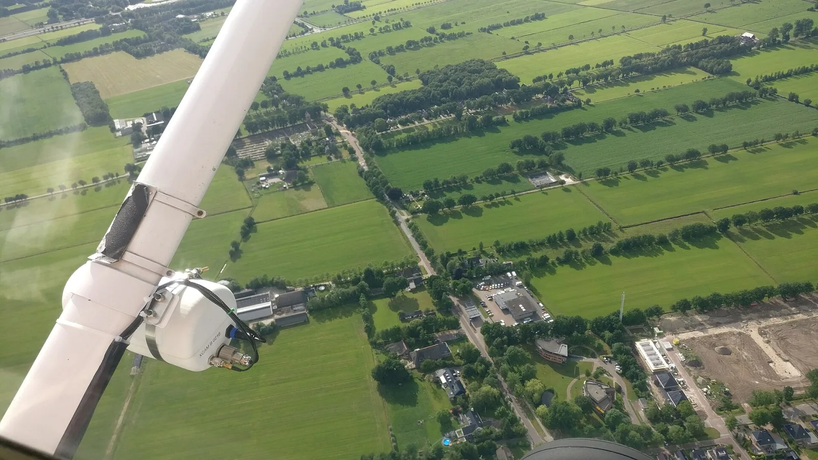Seen from the cockpit of a small plane, a white survey camera pod is fixed to the wing strut above a patchwork of fields and villages