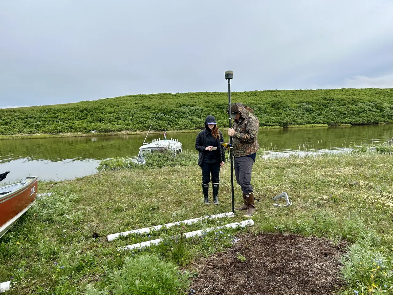Two people in rain gear stand on a grassy riverbank in Alaska, one holding a satellite positioning pole over a survey marker, with white measuring rods on the ground