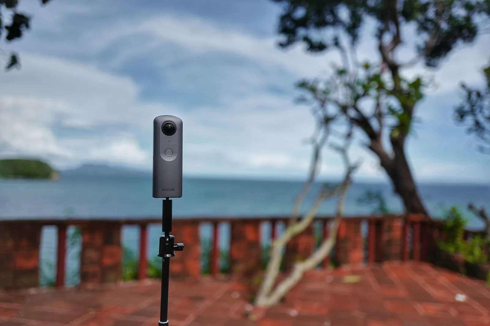 A slim 360 camera, a Ricoh Theta V, mounted on a thin tripod pole on a terrace overlooking the sea