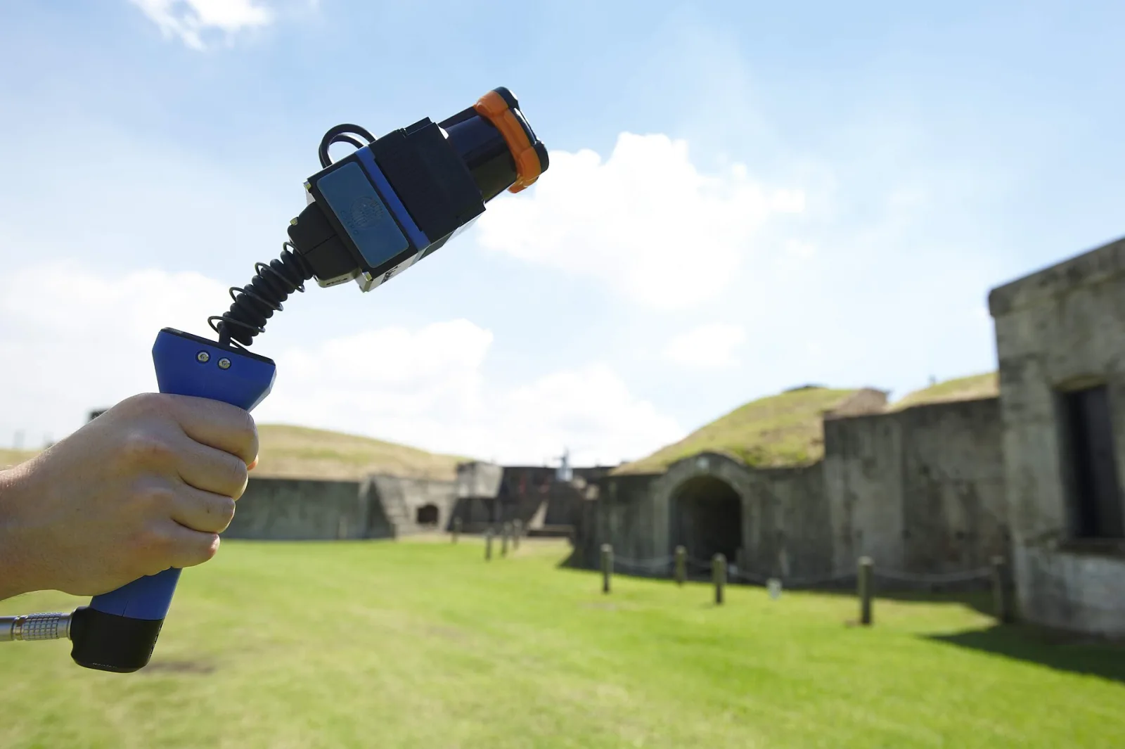 A hand holds up a handheld LiDAR scanner, a small rotating sensor on a spring above a blue handle, in front of the stone walls of an old fort
