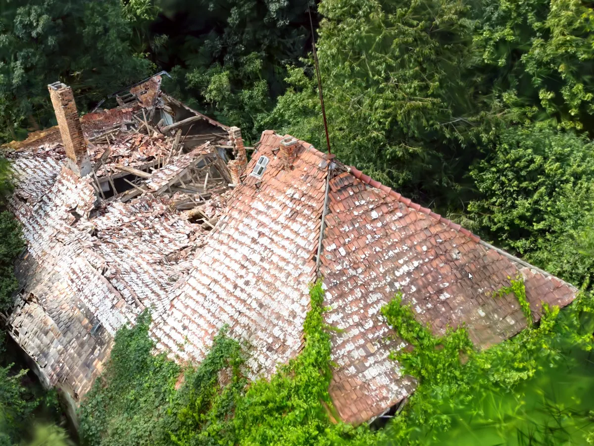 Seen from above, a house whose roof has partly collapsed, with broken tiles, exposed beams and a brick chimney, surrounded by trees, rendered from a Gaussian splat