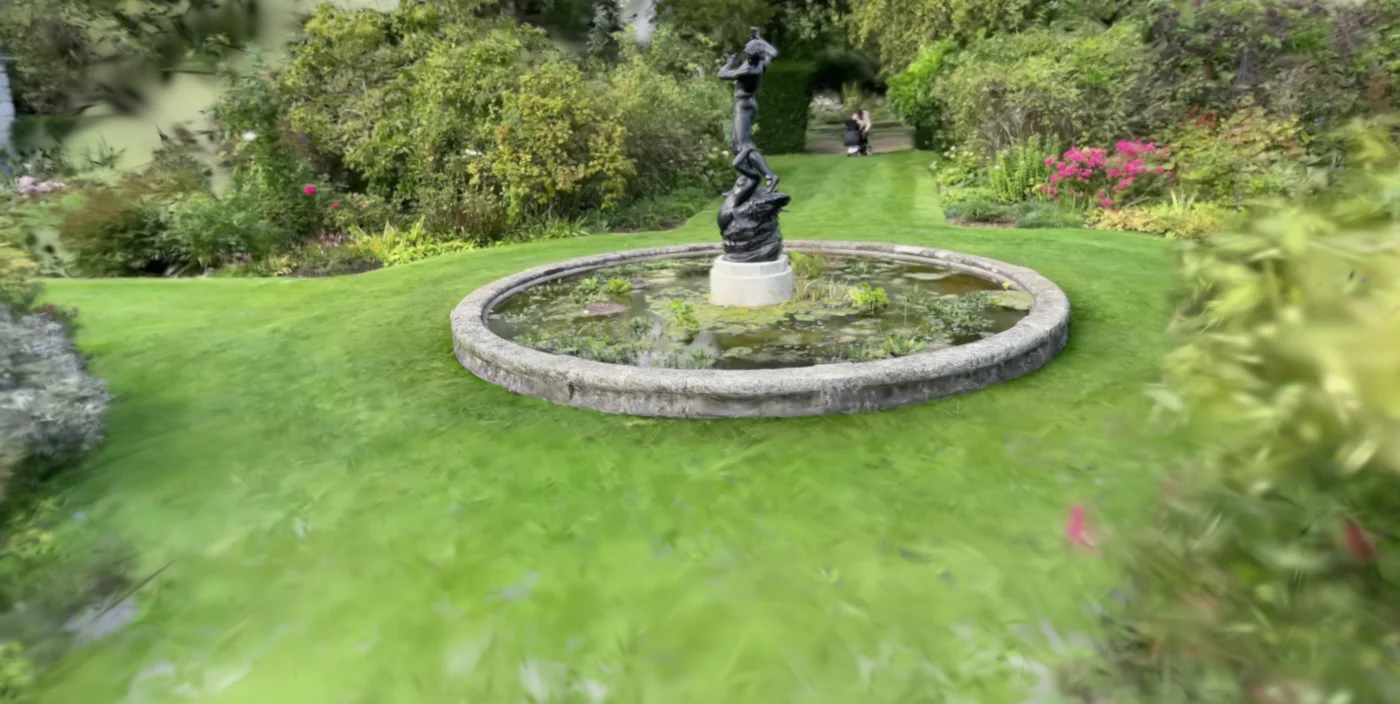 A garden with a round stone fountain and a bronze statue, on a lawn edged with flower beds, rendered from a Gaussian splat; the edges of the image are blurred